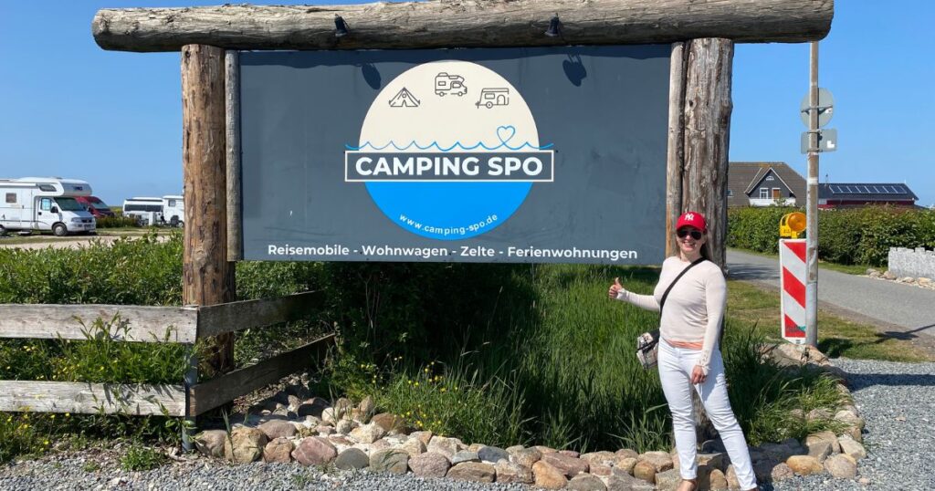 Blonde Frau steht vor dem Schild eines Camping Platzes